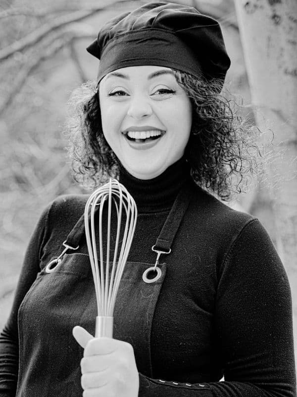 Pinar, the founder and chef of MsPinaQ, wearing a black chef's hat and apron while holding a wire whisk