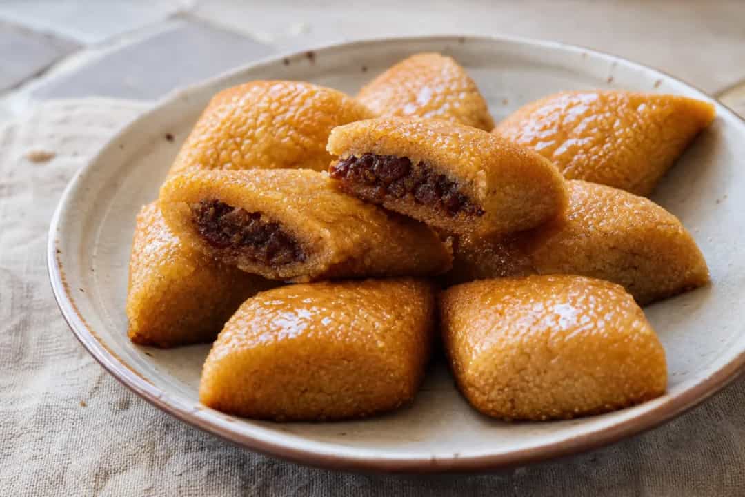 Golden-brown Algerian makroud pastries filled with sweet dates, drizzled with honey, served on a decorative plate.