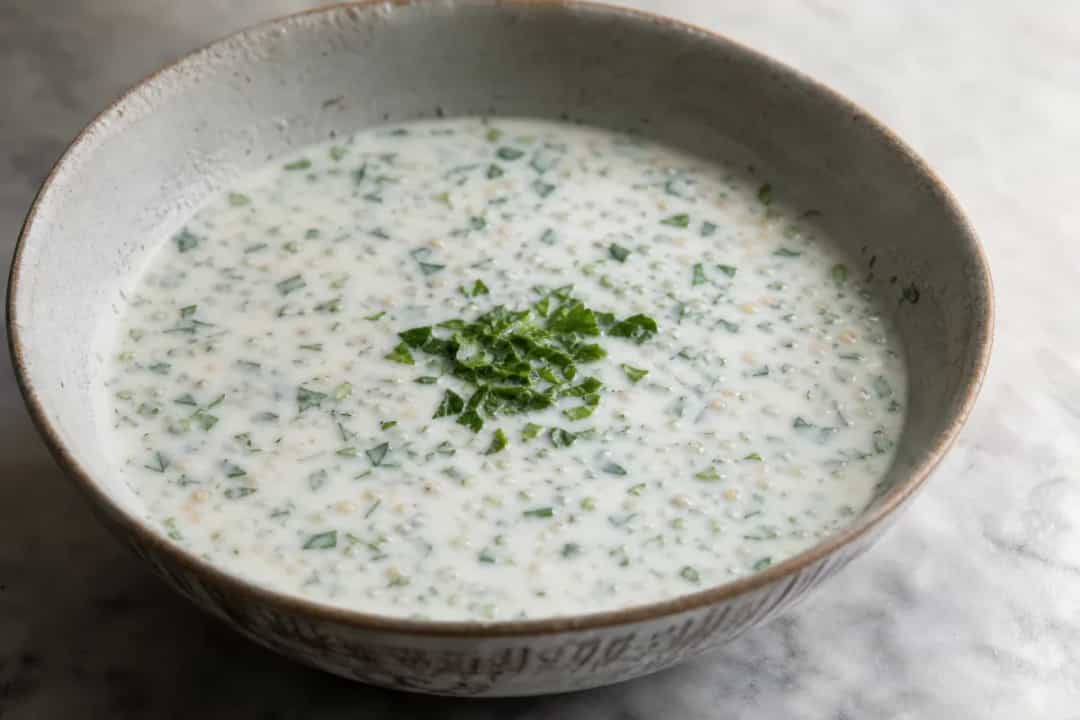 Creamy yogurt soup garnished with fresh herbs and wheat, served in a bowl.
