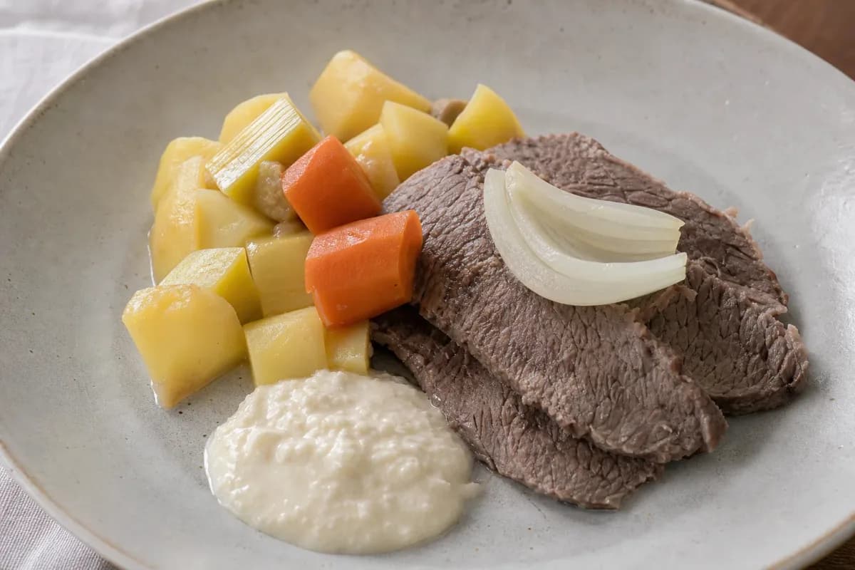 Tender slices of boiled beef with colorful root vegetables and creamy horseradish sauce elegantly plated.
