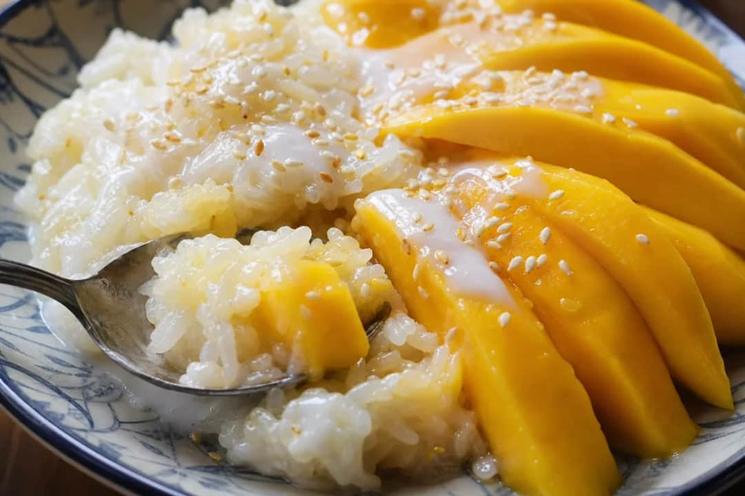 Plated Thai mango sticky rice topped with coconut sauce and fresh mango slices, garnished with sesame seeds.