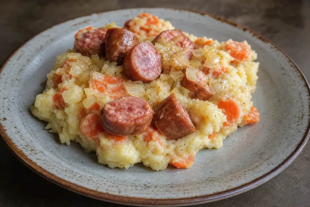 Creamy mashed potatoes and carrots with sliced sausage in a bowl, garnished with herbs.