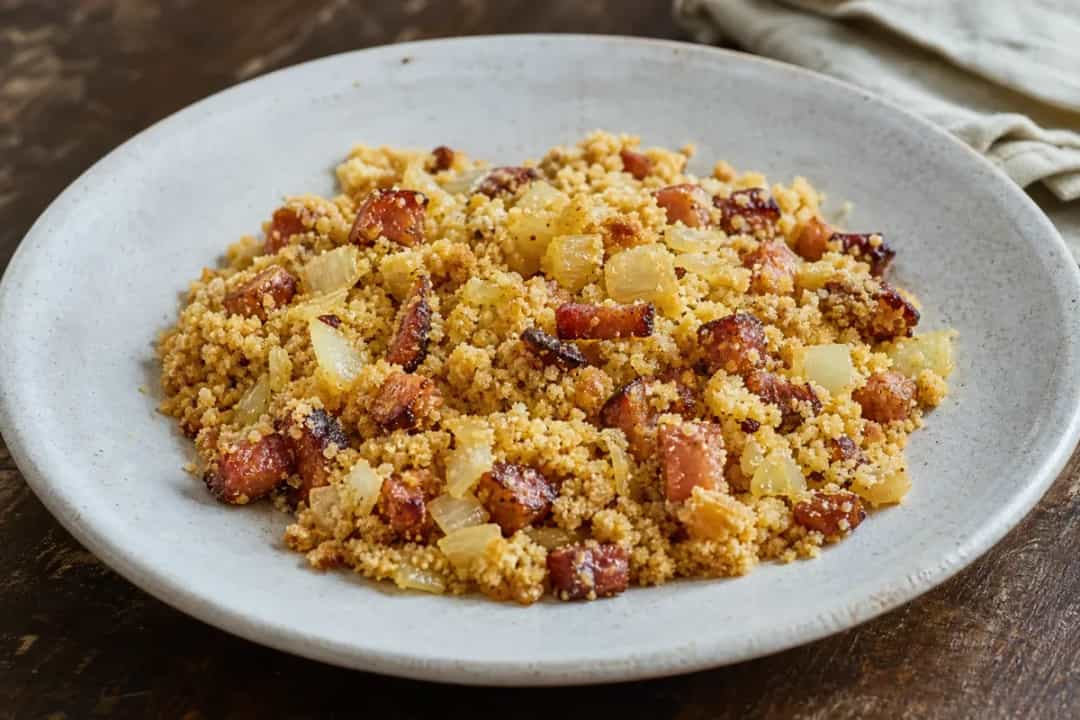 Golden brown farofa sprinkled with crispy bacon and sautéed onions on a white plate.