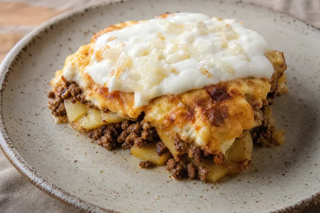 Layered moussaka with golden potatoes, minced meat, and creamy yogurt topping, served in a rustic dish.