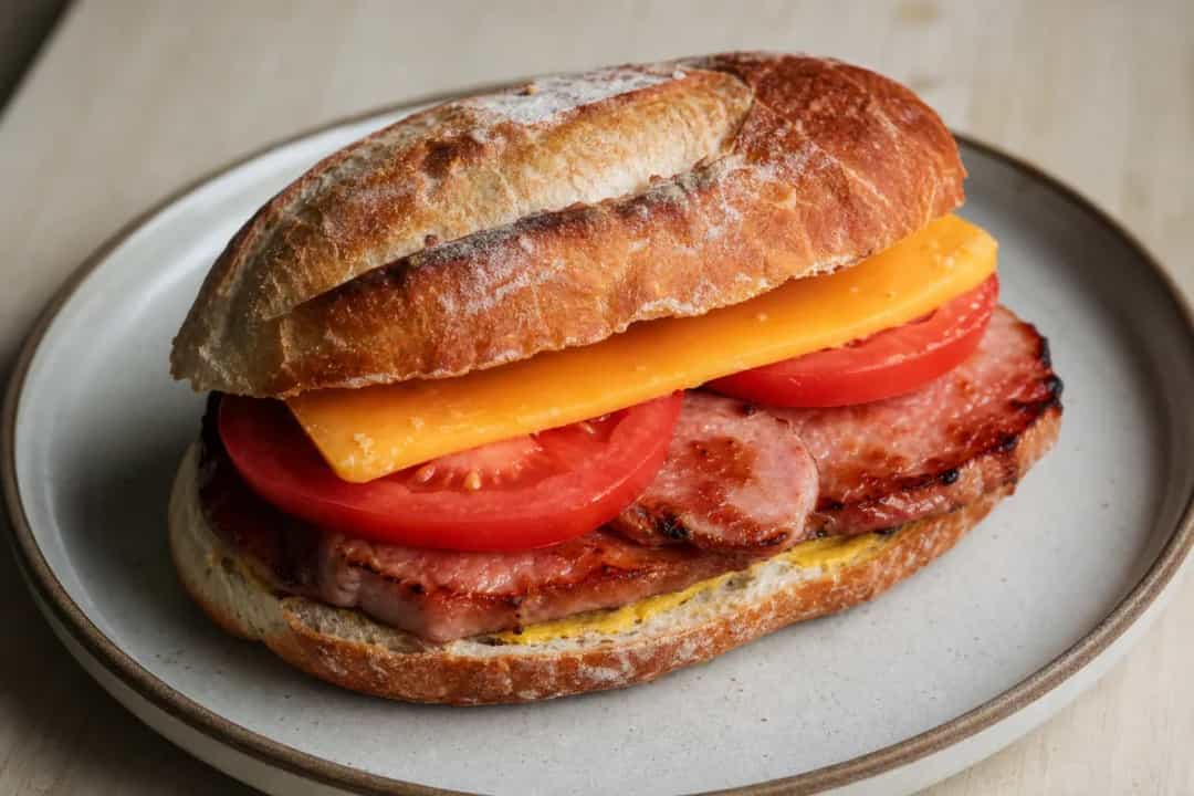A hearty sandwich with golden peameal bacon, mustard, and fresh lettuce on crusty bread, garnished with pickles.