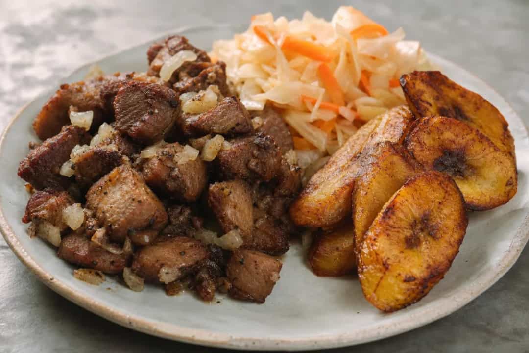 Crispy fried pork served with spicy Pikliz and golden fried plantains, garnished beautifully.