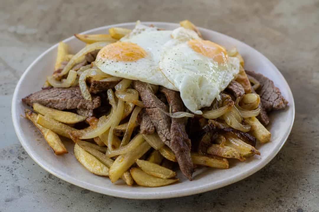 A vibrant plate of Chorrillana with golden fries, sliced beef, onions, and sunny-side-up eggs, garnished with parsley.
