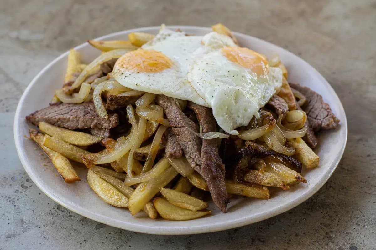 A vibrant plate of Chorrillana with golden fries, sliced beef, onions, and sunny-side-up eggs, garnished with parsley.