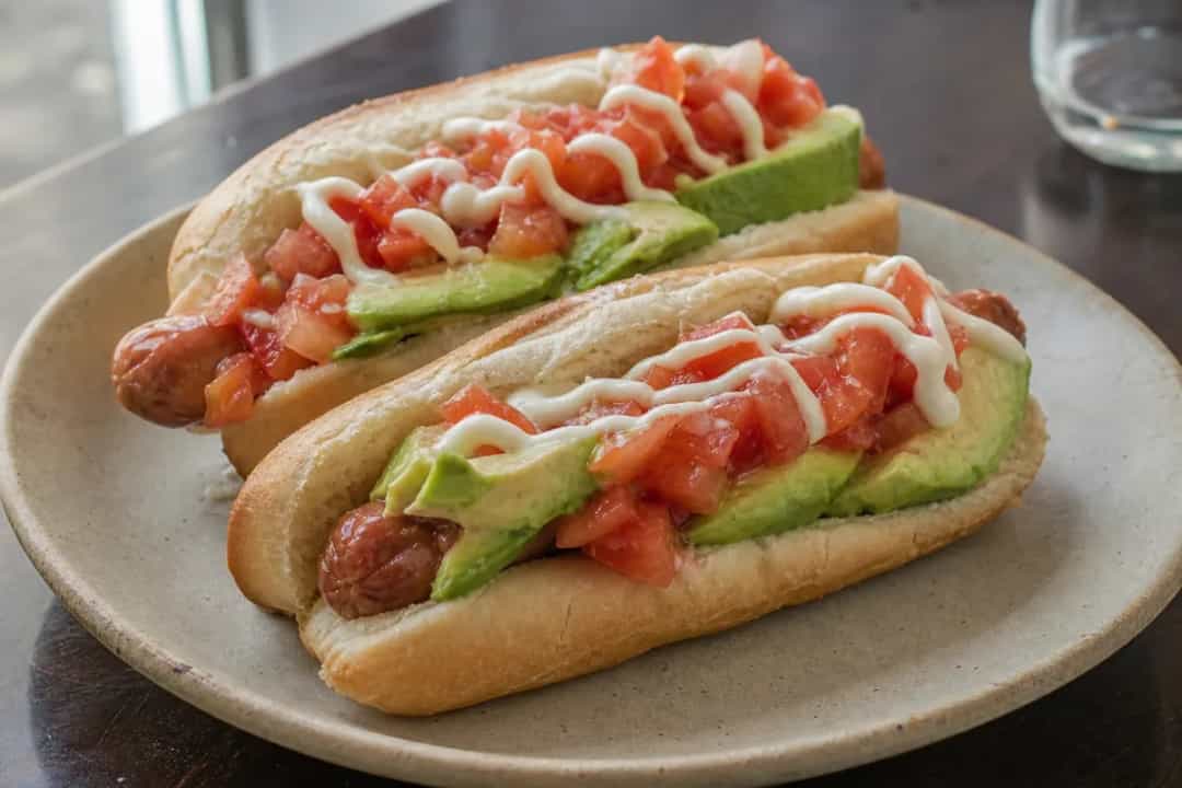 A colorful plate of Completo Italiano featuring a hot dog with creamy avocado, diced tomatoes, and mayonnaise garnished with parsley.