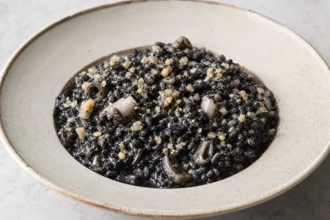 Plated black risotto garnished with cuttlefish, squid ink, and herbs.