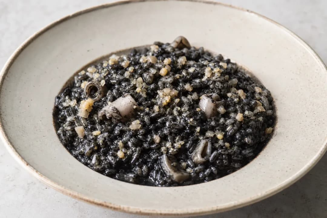 Plated black risotto garnished with cuttlefish, squid ink, and herbs.