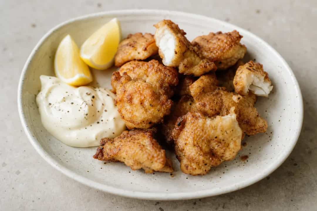 Golden fried fish bites served with creamy garlic sauce and lemon wedges, garnished with fresh herbs.
