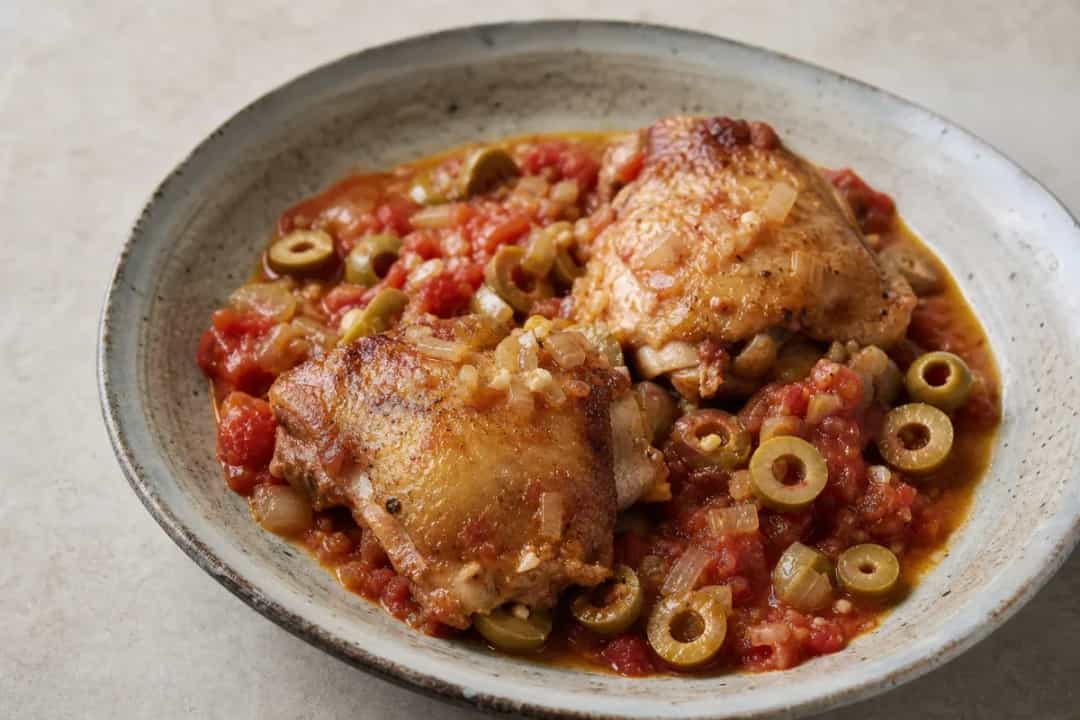 A beautifully plated chicken dish with vibrant tomatoes, green olives, and fresh herbs on a rustic background.