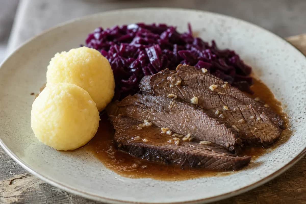 Plated German Sauerbraten with tender meat, vibrant red cabbage, and fluffy potato dumplings garnished with herbs.