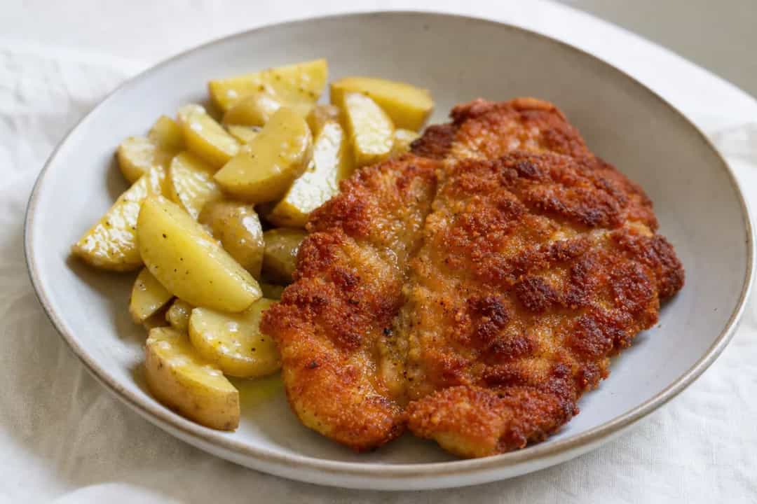 A plate of crispy schnitzel with a side of warm potato salad garnished with lemon slices and fresh herbs.