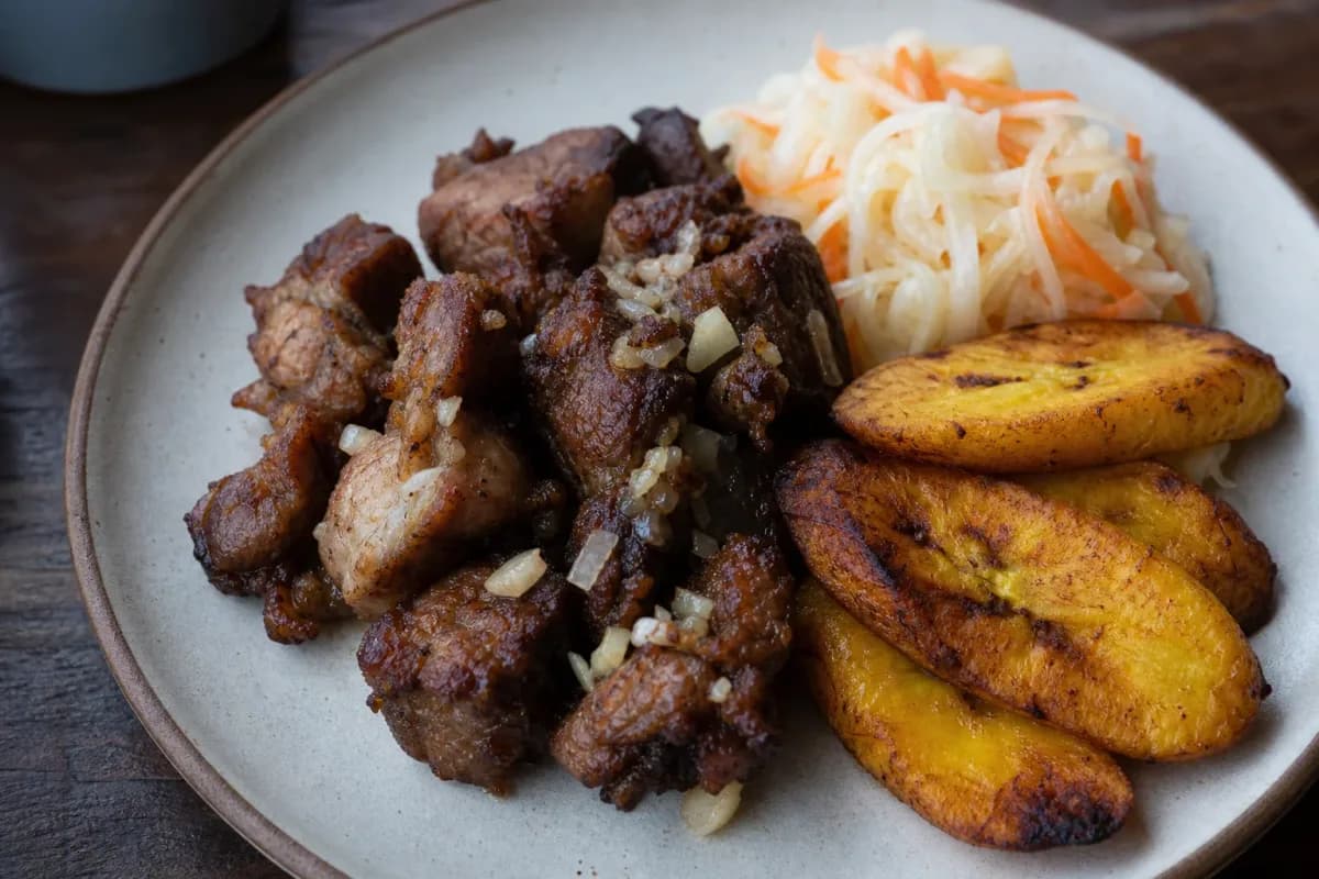Plated Haitian Griot with vibrant Pikliz and golden fried plantains, showcasing rich flavors and textures.