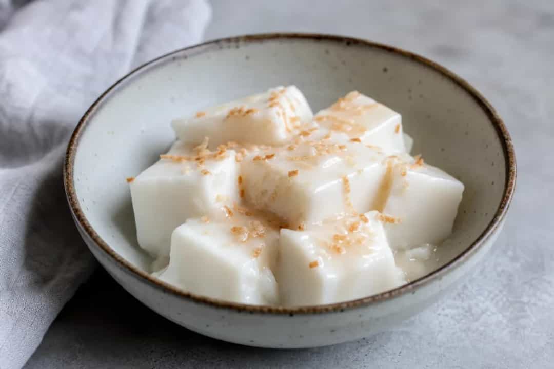 Creamy coconut pudding squares topped with shredded coconut and served in a dish.