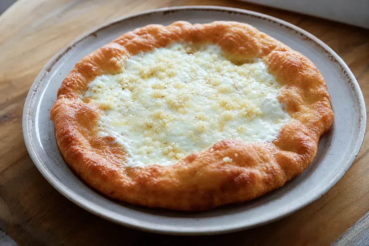 Golden-brown lángos topped with creamy sour cream, melted cheese, and garlic, garnished with herbs on a rustic plate.
