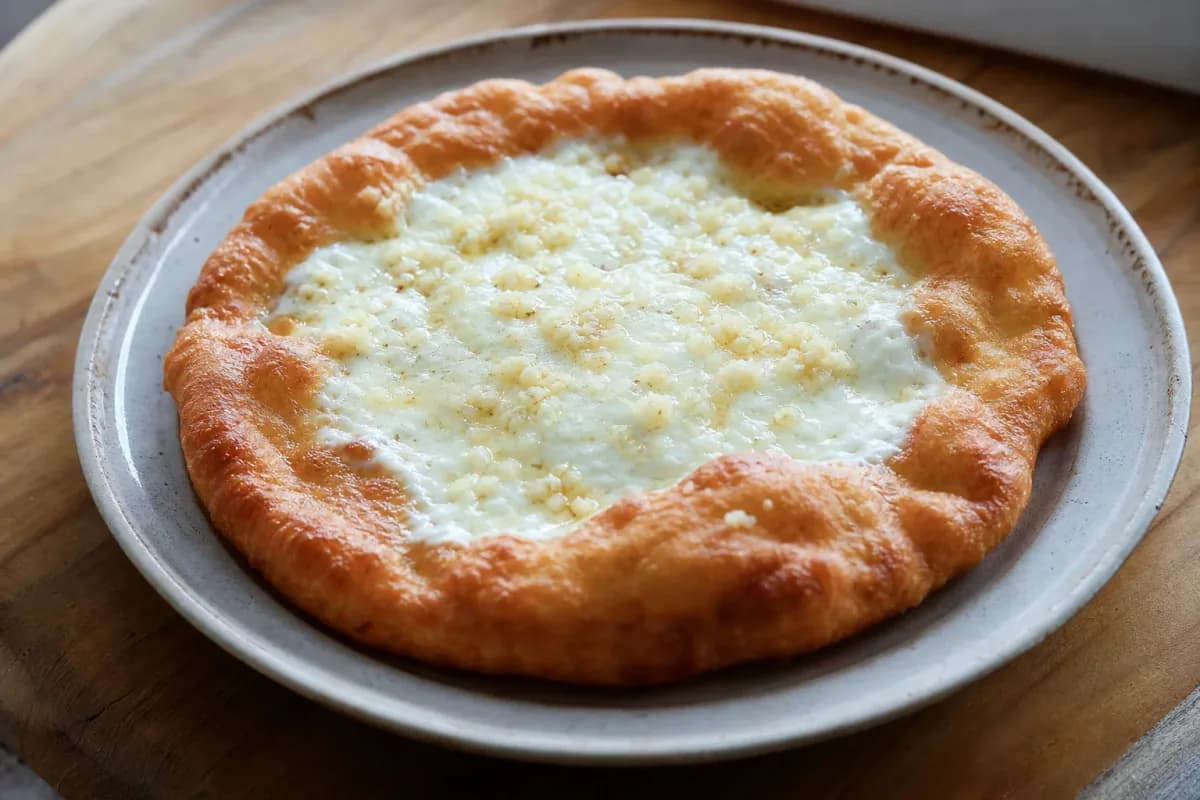 Golden-brown lángos topped with creamy sour cream, melted cheese, and garlic, garnished with herbs on a rustic plate.