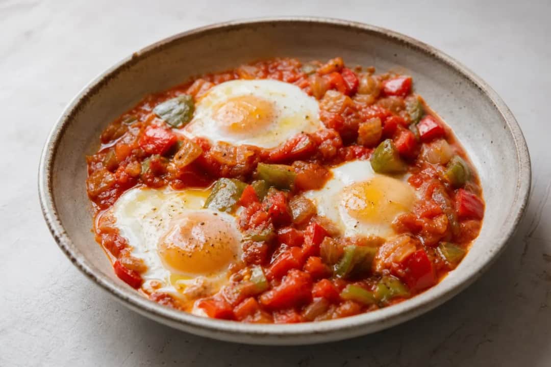 A vibrant shakshuka with poached eggs, red peppers, and tomatoes topped with fresh herbs in a skillet.