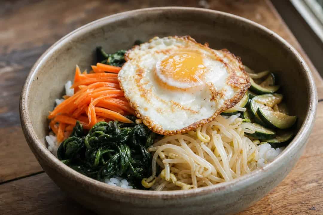 Colorful bibimbap bowl with sautéed vegetables, fried egg, and rice garnished with sesame seeds.