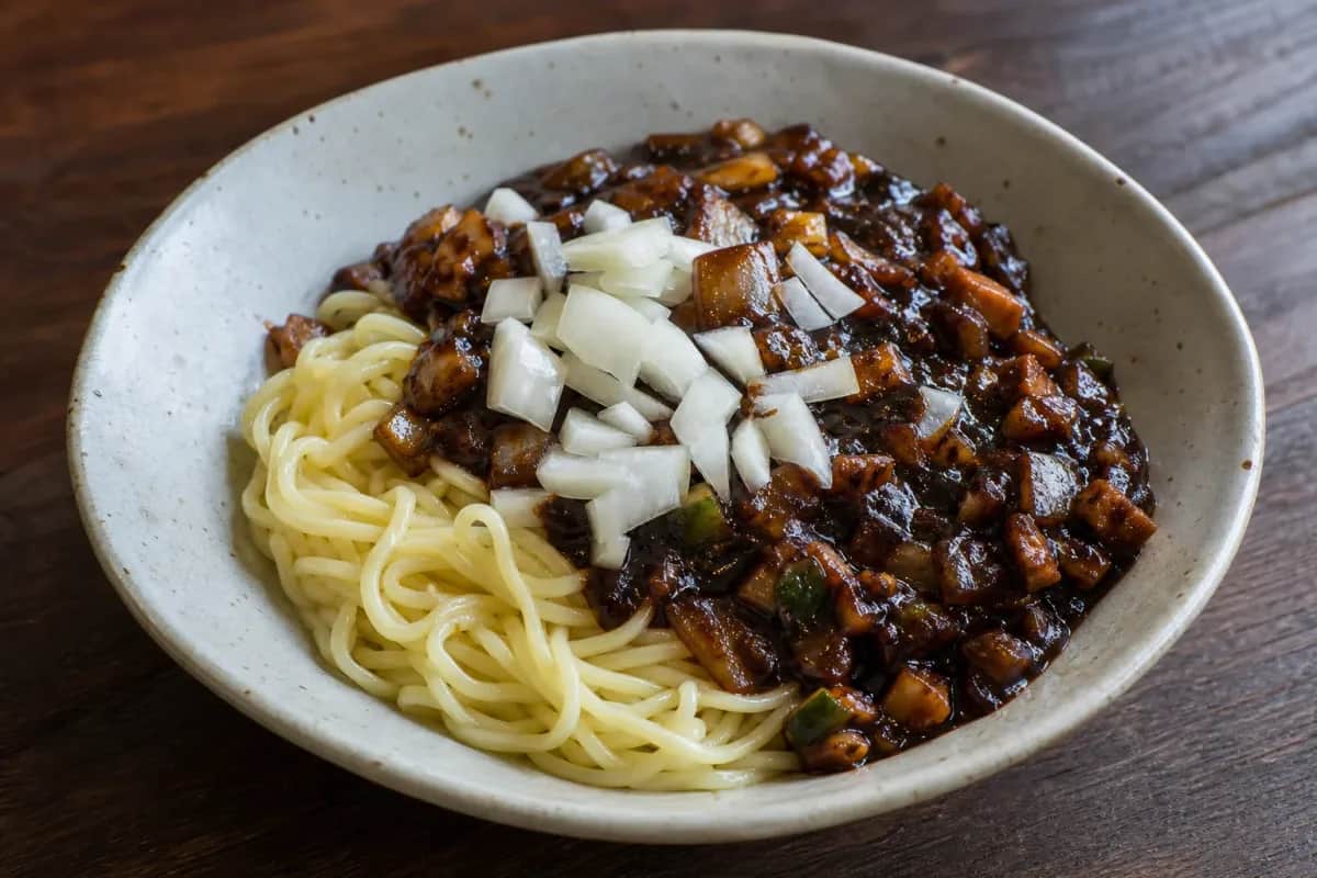 A bowl of Jajangmyeon featuring thick noodles covered in glossy black bean sauce, garnished with diced cucumbers and onions.