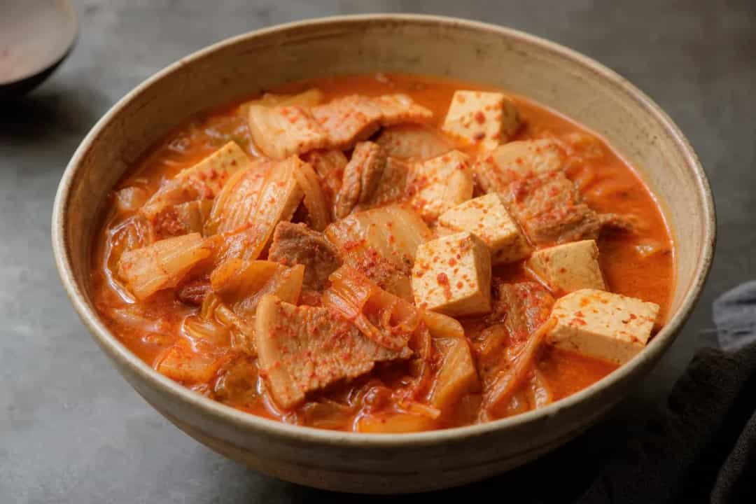 A steaming bowl of kimchi stew with tofu, garnished with green onions and chili, showcasing vibrant colors.