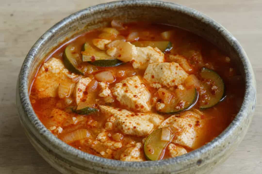 A bowl of Korean Sundubu Jjigae with soft tofu, vegetables, and a spicy broth, garnished with green onions.
