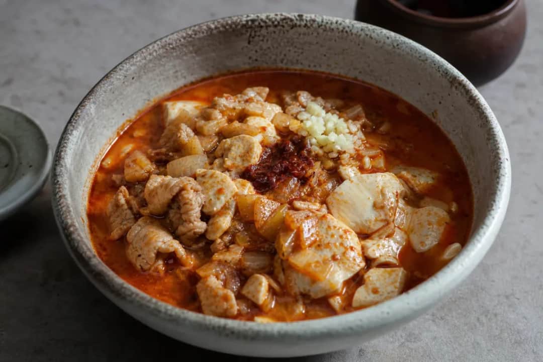 Bowl of Korean sundubu jjigae with soft tofu, vegetables, and spicy broth, garnished with green onions.