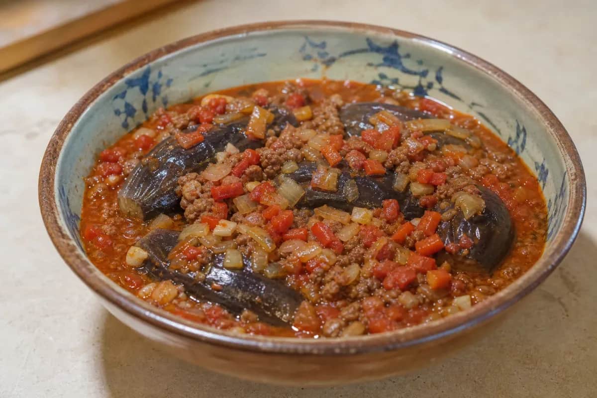 A steaming bowl of eggplant and meat stew with vibrant tomatoes and spices, garnished with fresh herbs.