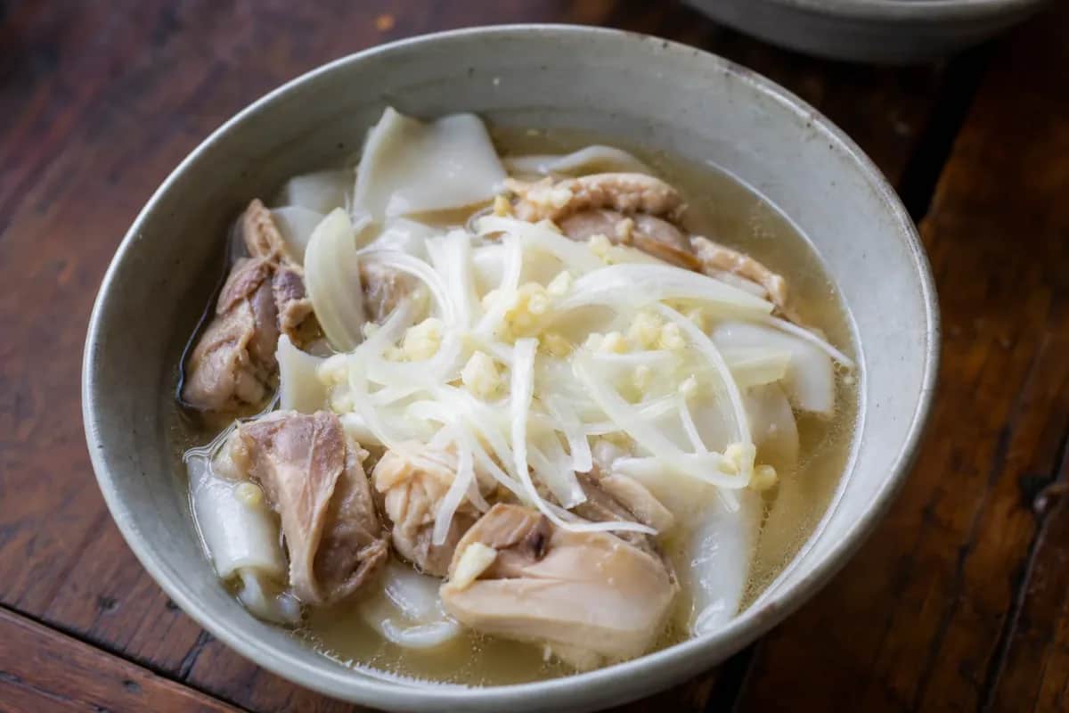 A bowl of Khao Piak Sen with smooth rice noodles, tender chicken, and fresh herbs garnished on top.