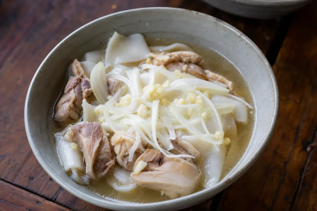 A bowl of Khao Piak Sen with smooth rice noodles, tender chicken, and fresh herbs garnished on top.