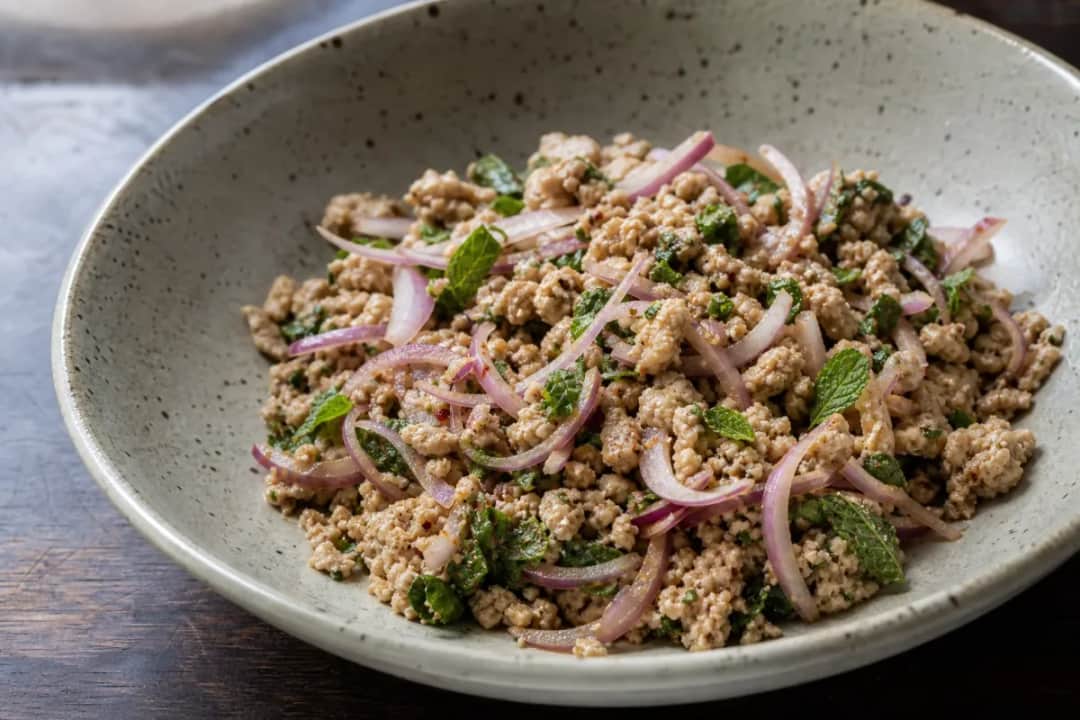 A colorful bowl of Laap featuring minced chicken, toasted rice, and fresh herbs garnished with lime.