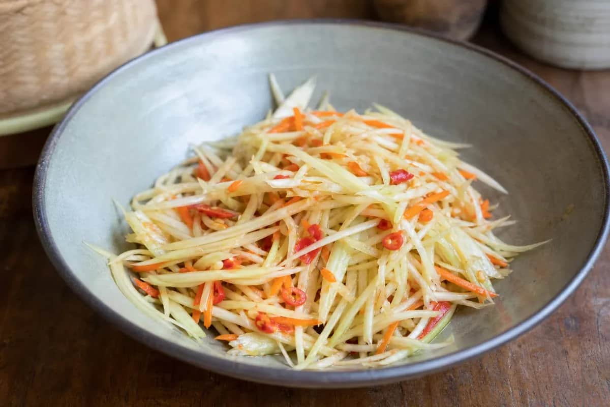 A colorful bowl of spicy green papaya salad topped with crushed peanuts and fresh herbs, showcasing vibrant textures and flavors.
