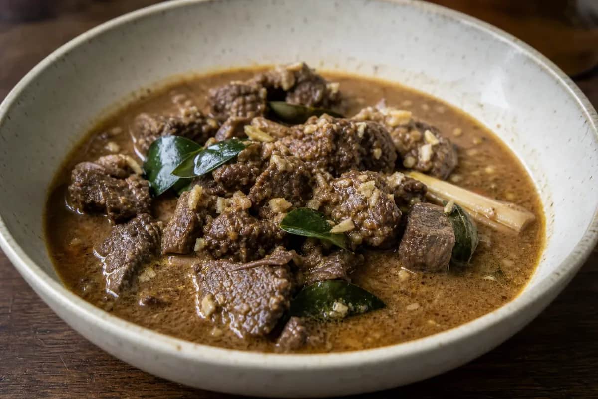 Rich beef rendang garnished with herbs, served in a bowl with coconut sauce and spices.