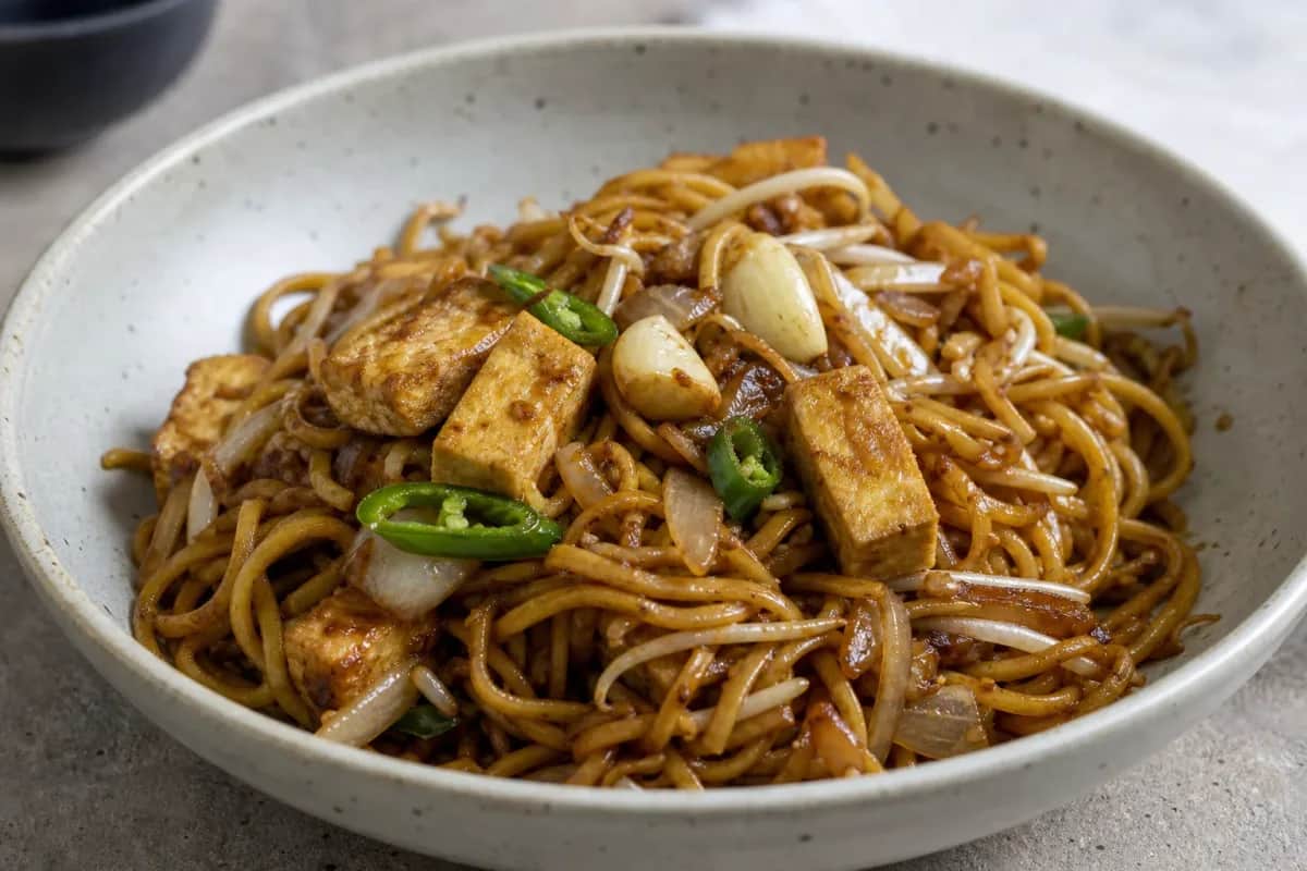 Colorful plate of mee goreng with stir-fried noodles, tofu, and vegetables garnished with chili sauce.