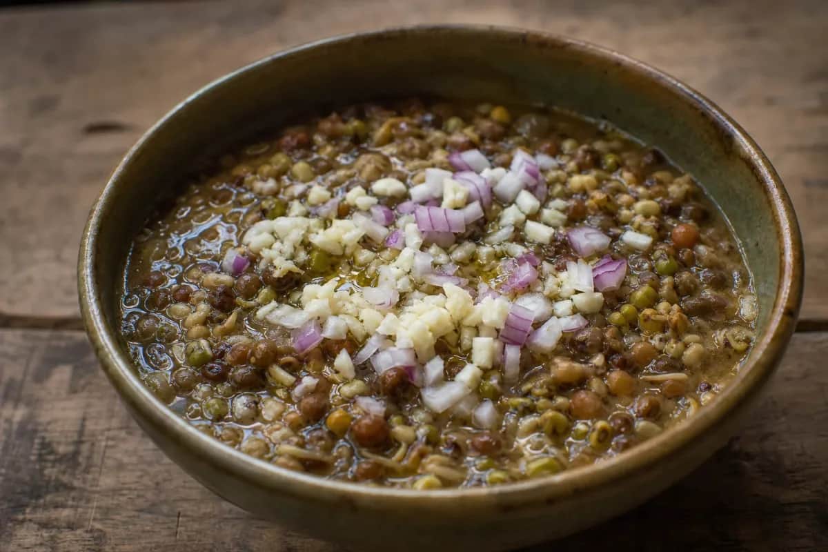 A bowl of thick, hearty Kwati soup garnished with fresh herbs and spices, showcasing mixed sprouted beans.