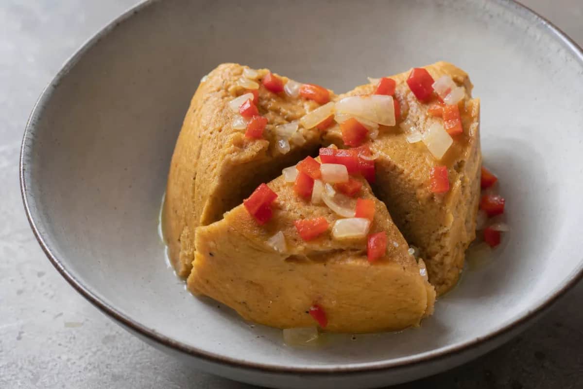 Smooth and colorful Nigerian Moi Moi served on a plate with vibrant pepper sauce and garnished with herbs.