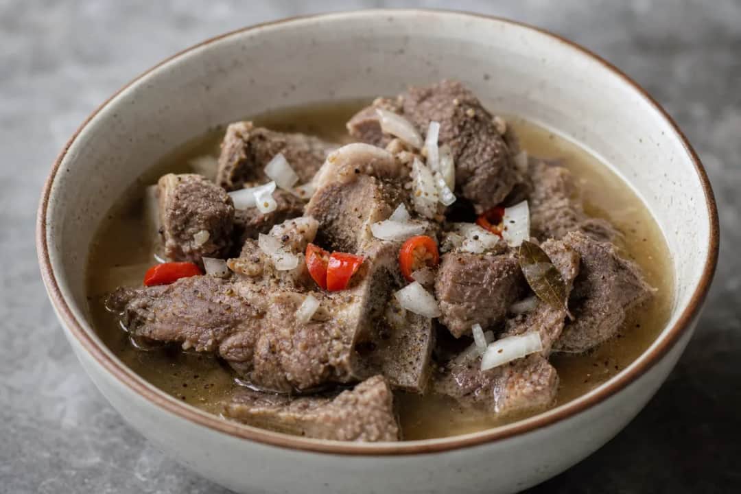 Bowl of Nigerian pepper soup with goat meat, herbs, and spices, garnished with green onions.