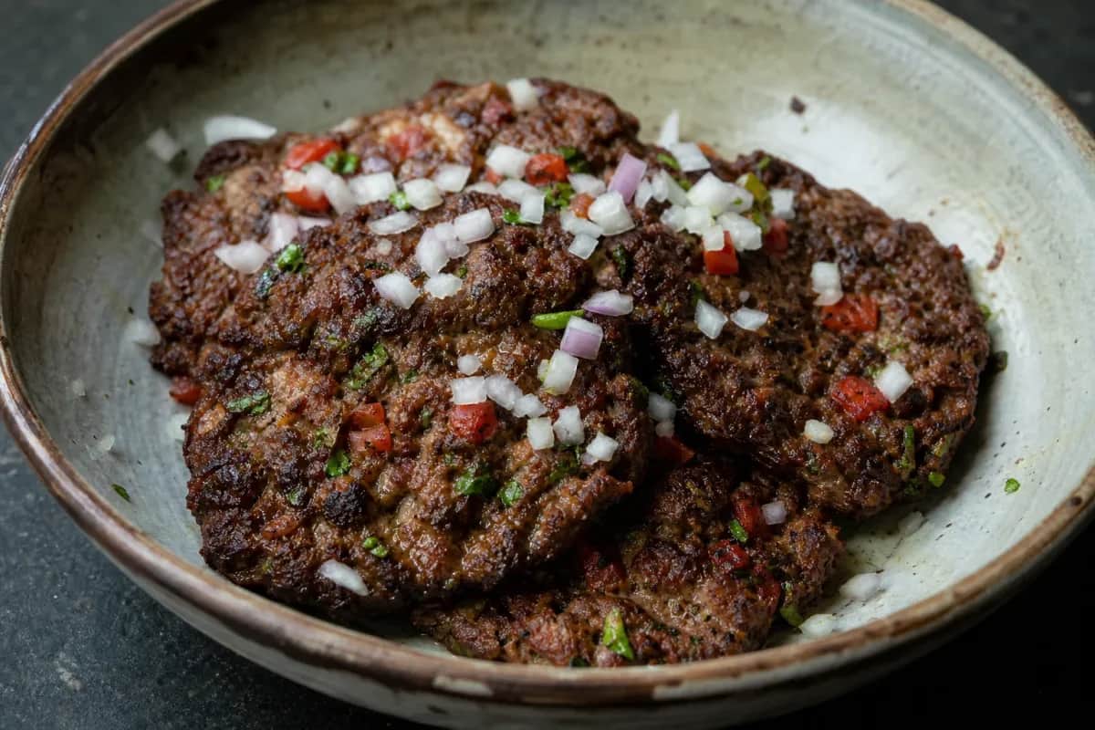 Golden-brown chapli kebabs garnished with fresh herbs and sliced tomatoes, served on a plate.