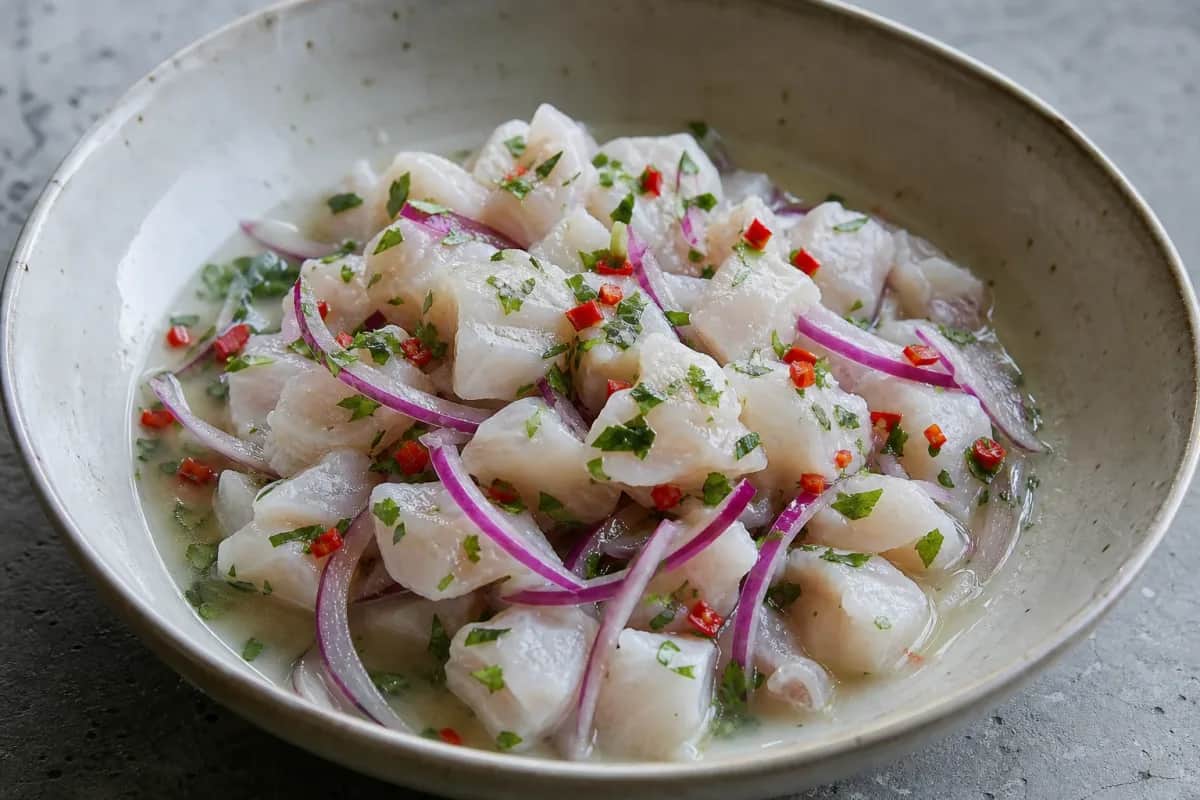 Plated ceviche with bright white fish, red onion slices, and green chili, garnished with lime wedges.