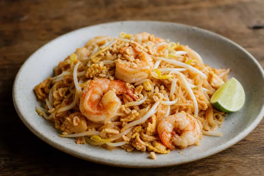 A vibrant plate of Pad Thai with shrimp, garnished with peanuts and lime wedges, showcasing colorful vegetables and glossy noodles.