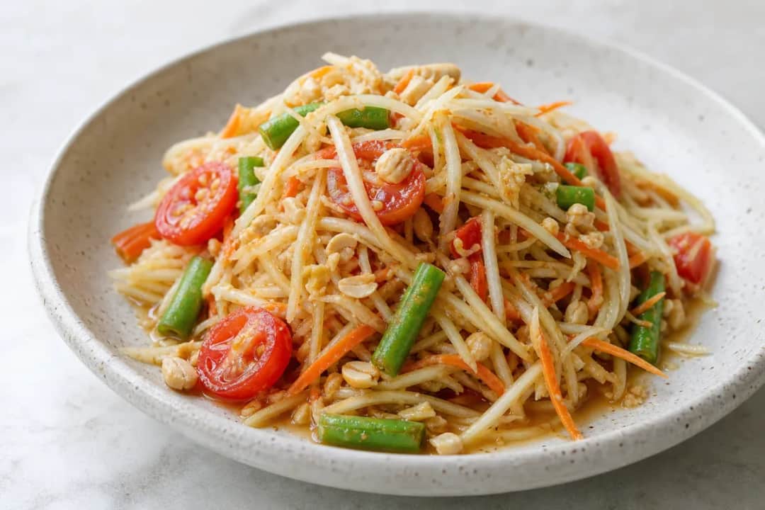 A vibrant plate of Thai Som Tam salad featuring shredded green papaya, tomatoes, peanuts, and a drizzle of dressing, garnished with fresh herbs.